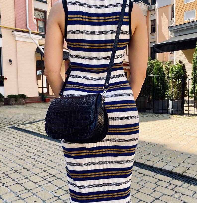 May include: A black, textured leather crossbody bag with a flap closure and a silver-tone chain strap. The bag is worn over a striped dress with white, navy, and gold horizontal stripes. The dress has a sleeveless design.