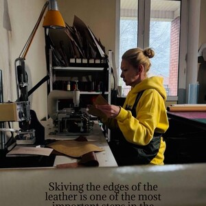 May include: A person in a yellow hoodie working with leather in a workshop. The text in the image reads: "Skiving the edges of the leather is one of the most important steps in the making process - not a single bag is made without it."