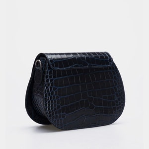 May include: A navy blue, semi-circular crossbody bag with a crocodile skin texture. The bag has a flap closure and a silver-toned metal clasp. The strap is attached to the sides of the bag. The bag is set against a white background.