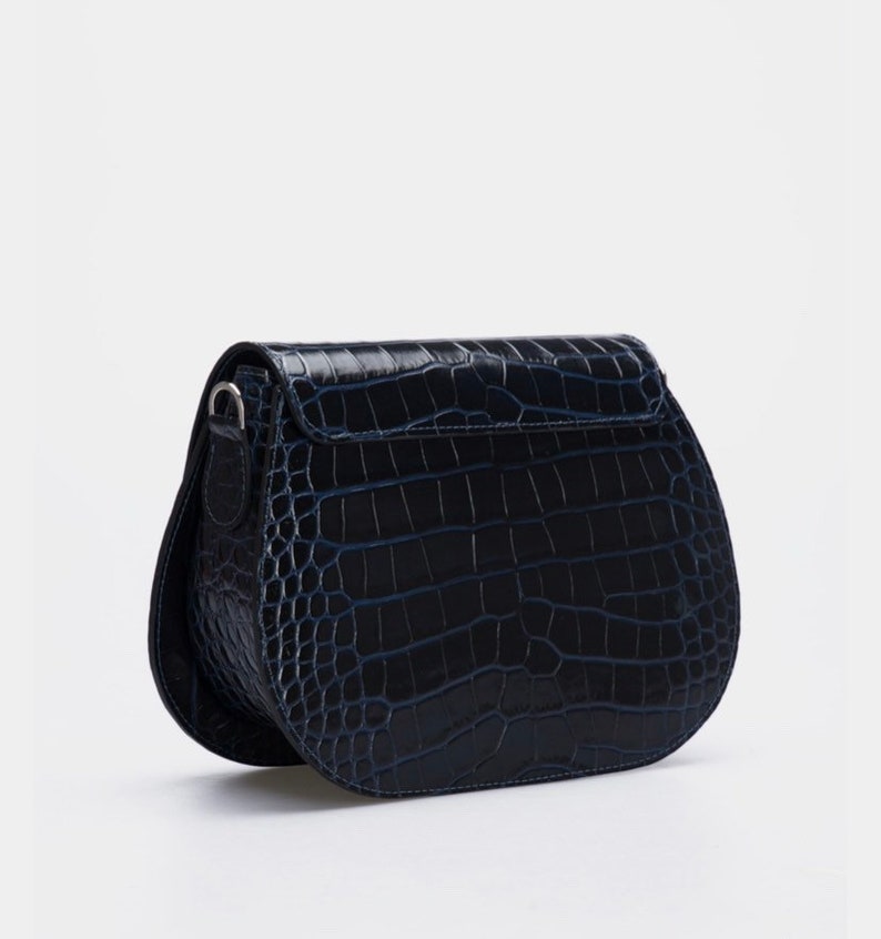 May include: A navy blue, semi-circular crossbody bag with a crocodile skin texture. The bag has a flap closure and a silver-toned metal clasp. The strap is attached to the sides of the bag. The bag is set against a white background.
