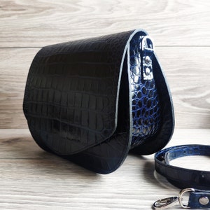 May include: A black leather crossbody bag with a crocodile texture. The bag has a curved shape with a flap closure and a blue interior. A matching strap and silver hardware are also visible.