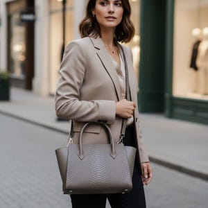 Peut inclure: Un sac à main taupe doté d'un panneau central texturé à motif peau de serpent, de deux poignées supérieures et d'une bandoulière amovible. Le sac présente une silhouette structurée avec des panneaux latéraux lisses et des attaches argentées.