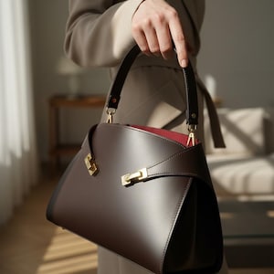 May include: A structured dark brown handbag featuring a smooth finish, a single top handle, and gold-toned metal hardware accents. The bag has a distinct curved silhouette with a visible red interior lining and a secure clasp closure mechanism.