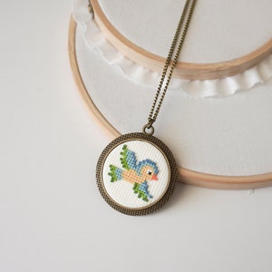May include: A blue bird cross-stitch design in a round bronze frame on a chain necklace.