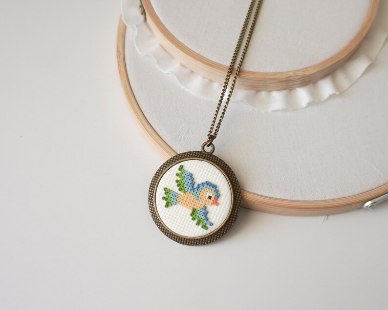 May include: A blue bird cross-stitch design in a round bronze frame on a chain necklace.