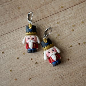 Può includere: Un paio di orecchini pendenti festivi con figure di schiaccianoci in miniatura. Ogni orecchino ha un cappello nero con finiture dorate, una giacca rossa e una barba bianca. Gli orecchini sono realizzati in pasta polimerica.
