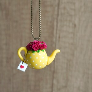 Pode incluir: Um colar de chaleira amarela com bolinhas brancas e um buquê de flores rosa no topo. A chaleira tem uma pequena etiqueta com um coração vermelho.
