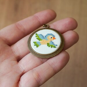 May include: A small, round, bronze-colored pendant with a white background and a blue and green embroidered bird design.