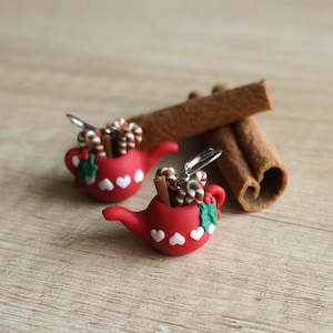 Könnte beinhalten: Rote Teekannen-Ohrringe mit weißen Herzen, gefüllt mit Zimtstangen und Zuckerstangen. Die Ohrringe liegen auf einer Holzoberfläche mit Zimtstangen.