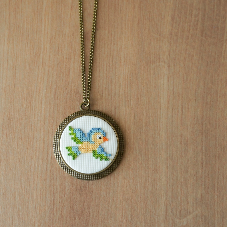 May include: A bronze pendant necklace with a white circular canvas featuring a blue and yellow embroidered bird.