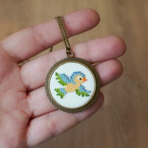 May include: A bronze pendant necklace with a round, white canvas featuring a cross-stitch design of a blue and yellow bird with green wings.