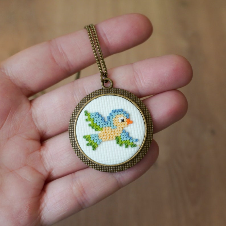 May include: A bronze pendant necklace with a round, white canvas featuring a cross-stitch design of a blue and yellow bird with green wings.