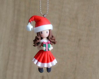 May include: A handmade Christmas ornament featuring a girl wearing a red and white Santa hat, a red dress, and holding a green gift box.