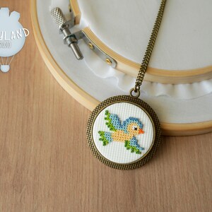 May include: A blue bird cross-stitch design on a white background, framed in a bronze-colored circular pendant. The pendant is attached to a gold chain necklace.