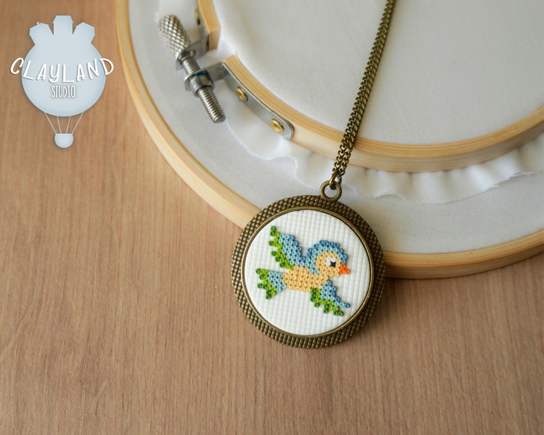 May include: A blue bird cross-stitch design on a white background, framed in a bronze-colored circular pendant. The pendant is attached to a gold chain necklace.