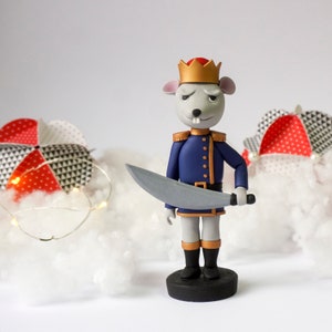 Pode incluir: Uma figurinha de rato cinza vestida de soldado, com um uniforme azul e dourado, uma coroa vermelha e dourada, e segurando uma espada prateada. A figurinha está em pé sobre uma base preta.