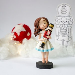 Może przedstawiać: Porcelanowa figurka lalki przedstawiająca młodą dziewczynkę w białej sukience trzymającą figurkę czerwonego i złotego działka. Lalka ma brązowe włosy i nosi niebieską wstążkę wokół talii. Działek ma złotą koronę i czerwony i złoty mundur. Lalka i działek stoją na czarnej podstawie.