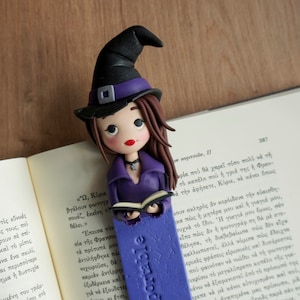 May include: Purple and black witch bookmark with a book in her hand. The bookmark is made of polymer clay and has a purple base with the word "portable" written in white.