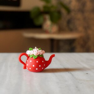 Könnte beinhalten: Eine rote Teekanne mit weißen Punkten und einem Blumendesign auf dem Deckel. Die Teekanne ist aus Keramik und hat einen Ausguss und einen Griff.