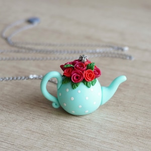 Pode incluir: Um pingente de chaleira azul turquesa com bolinhas brancas e um buquê de rosas vermelhas no topo.