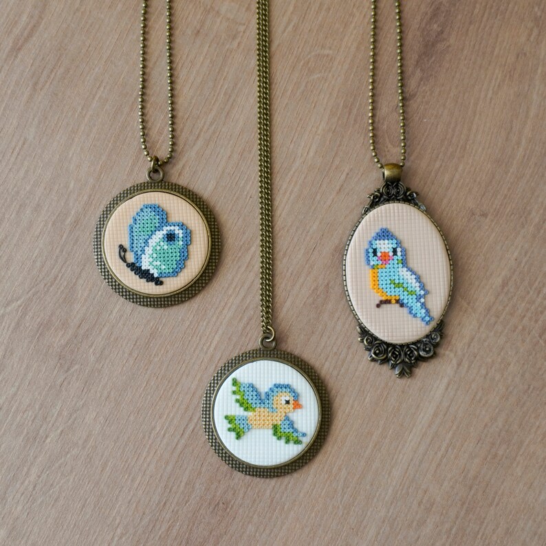 May include: Three necklaces with cross-stitch embroidery on round metal pendants. The pendants feature a blue butterfly, a blue bird, and a blue bird in flight. The necklaces are on a wooden surface.
