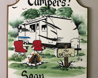 May include: A white wooden sign with a hand-painted illustration of a camper, two red chairs, and a campfire. The sign reads "Happy Campers!" and "Sean and Ginni" with a sign that says "The Hiers'" in the foreground.
