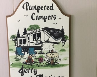 May include: A wooden sign with a brown border and a white background. The sign features a cartoon illustration of a camper van, two chairs, a campfire, and a dog. The text on the sign reads "Pampered Campers", "Reflection", "The Spinas", "Ginger", "Jerry and Marianne".
