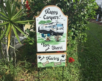 May include: A wooden sign with a white background and brown trim. The sign has a curved top and is painted with a picture of a motorhome, two chairs, a campfire, and a sign that says "Camping Sweet Home". The sign also has the text "Happy Campers!" at the top, "Tom and Sherri" in the middle, and "and Harley too!" at the bottom. The sign is on a metal stand.