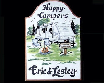 May include: A wooden sign with a painted image of a camper van parked in a campsite with a campfire and two chairs. The sign has the text "Happy Campers" at the top and "Eric & Lesley" at the bottom.