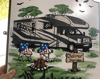 May include: A white wooden sign with a hand-painted illustration of a motorhome, two camping chairs, and a campfire. The sign reads "Life is Good!" and "The Johnson Family" with a sign that says "Oldsman Florida" in the foreground.