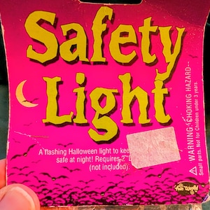 May include: A bright pink cardboard packaging for a Flashing Safety Light. The text reads Flashing Safety Light with a crescent moon graphic and a swarm of black bats at the bottom. It includes a choking hazard warning and requires 2 D batteries.
