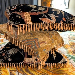 May include: A stack of three square throw pillows featuring intricate woven tapestry patterns in black, gold, and muted earth tones. Each pillow is finished with a decorative fringe trim consisting of small, dangling wooden bead accents.