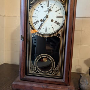 May include: Antique wooden wall clock with a rectangular frame. The clock face is white with black Roman numerals and the word "CORNE" printed in the center. The clock has a pendulum visible through a glass panel, and decorative gold accents.