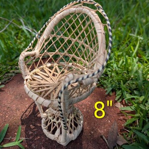 May include: A small, handcrafted wicker chair, approximately 8 inches tall. The chair features a woven seat and backrest, with a decorative rim of black and white stripes. The chair is made of natural-colored wicker and is set against a backdrop of green grass.