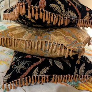May include: A stack of three square throw pillows featuring woven fabric with botanical patterns. The top and bottom pillows are black, while the middle pillow is gold. Each pillow is finished with a decorative fringe of small wooden beads.