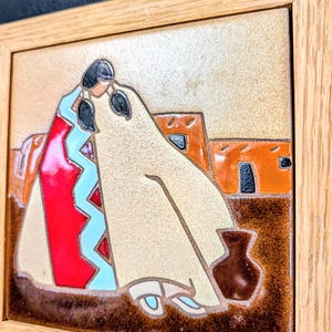 May include: A square ceramic tile framed in light wood featuring a raised relief design of two figures wrapped in blankets. The figures are depicted in red, light blue, and cream colors against a background of brown earth and orange adobe structures.
