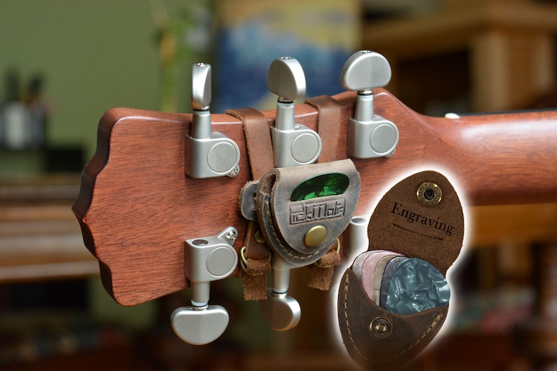 K&ouml;nnte beinhalten: Nahaufnahme einer Gitarrenkopfplatte mit silbernen Stimmwirbeln und einem braunen Leder-Plektrumhalter. Der Plektrumhalter ist mit Lederriemen befestigt und enth&auml;lt Gitarren-Plektren. Der Text "Engraving" ist sichtbar.