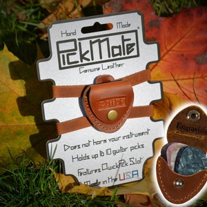 K&ouml;nnte beinhalten: Ein brauner PickMate Gitarrenplektrumhalter aus Leder mit dem Text "PickMate" und "Genuine Leather". Der Halter ist an einer wei&szlig;en Karte mit dem Text "Does not harm your instrument" und "Made in the USA" befestigt. Der Halter kann bis zu 10 Gitarrenplektren aufnehmen.