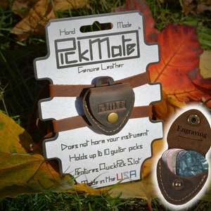 K&ouml;nnte beinhalten: Ein brauner PickMate Gitarrenplektrumhalter aus Leder mit Druckknopfverschluss, der mehrere Gitarrenplektren enth&auml;lt. Auf der Verpackung steht "Hand Made PickMate Genuine Leather" und "Made in the USA". Der Halter hat einen QuickPick-Schlitz und fasst bis zu 10 Plektren.