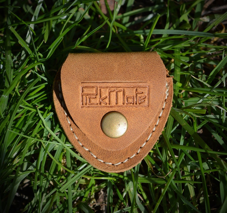 K&ouml;nnte beinhalten: Kleine, hellbraune Ledertasche mit Messingknopfverschluss. Die Tasche ist mit wei&szlig;em Faden gen&auml;ht und tr&auml;gt den gepr&auml;gten Text "RekMate". Sie liegt auf gr&uuml;nem Gras.