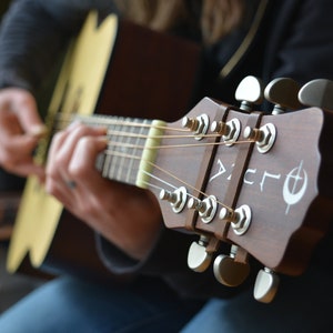 K&ouml;nnte beinhalten: Nahaufnahme der Kopfplatte einer braunen Akustikgitarre mit Stimmwirbeln. Auf der Kopfplatte steht das Wort "Lucia" und ein wei&szlig;es Symbol. Die Gitarrensaiten sind sichtbar, und eine Hand zupft die Saiten.