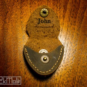 K&ouml;nnte beinhalten: Offener Gitarrenplektrumhalter aus braunem Leder mit dem Namen "John" auf der Klappe. Der Halter hat eine dunkelgraue Tasche und einen Druckknopfverschluss. Der Plektrumhalter liegt auf einer Holzoberfl&auml;che.