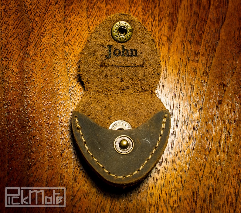 K&ouml;nnte beinhalten: Offener Gitarrenplektrumhalter aus braunem Leder mit dem Namen "John" auf der Klappe. Der Halter hat eine dunkelgraue Tasche und einen Druckknopfverschluss. Der Plektrumhalter liegt auf einer Holzoberfl&auml;che.