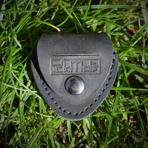 K&ouml;nnte beinhalten: Ein schwarzer Leder-Plektrumhalter mit dem gepr&auml;gten Text "PickMate" auf der Vorderseite. Der Halter wird mit einem silbernen Druckknopf verschlossen und ist von gr&uuml;nem Gras umgeben.
