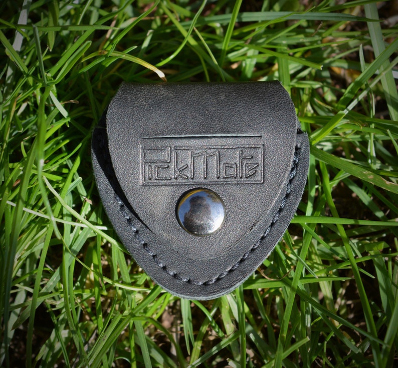 K&ouml;nnte beinhalten: Ein schwarzer Leder-Plektrumhalter mit dem gepr&auml;gten Text "PickMate" auf der Vorderseite. Der Halter wird mit einem silbernen Druckknopf verschlossen und ist von gr&uuml;nem Gras umgeben.