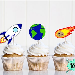 以下が含まれることがあります： 宇宙をテーマにした飾り付けがされたカップケーキが3つあります。左のカップケーキには青と白のロケット、真ん中のカップケーキには青と緑の地球、右のカップケーキには黄色の炎がついた茶色の彗星が飾られています。