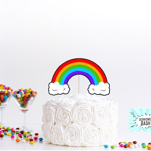 May include: A white cake with a rainbow cake topper. The rainbow is red, orange, yellow, green, blue, indigo, and violet. The cake is decorated with white frosting and has a cloud-like design on the sides. The cake topper is on a stick and has a white cloud on each side of the rainbow.