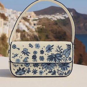 K&ouml;nnte beinhalten: Eine cremefarbene Handtasche mit Klappenverschluss und Tragegriff. Die Tasche ist mit einem detaillierten Blumenmuster in Blaut&ouml;nen verziert. Die Kanten und der Griff sind dunkelblau eingefasst. Im Hintergrund eine K&uuml;stenlandschaft.