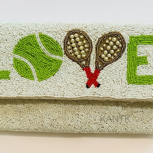 Puede incluir: Bolso de mano blanco con cuentas con la palabra "LOVE" en cuentas verdes. Una pelota de tenis verde y dos raquetas de tenis están representadas. Las raquetas son marrones con cuentas perladas y detalles rojos.