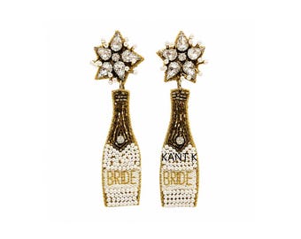 May include: A pair of champagne bottle earrings with a starburst design at the top. The bottles are embellished with gold and white beads, with the word "BRIDE" spelled out in gold letters. The earrings are designed for a wedding.